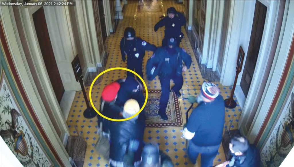 Police run at a group of men in the capitol. A man with a red cap wraps his arms around an officer